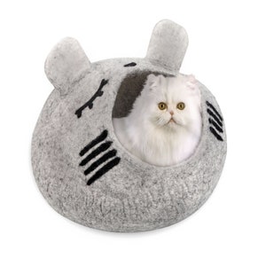 以下が含まれることがあります： 黒い刺繍の目とひげのある猫の顔の形をした灰色のフェルト製の猫用ベッド。白いペルシャ猫がベッドの中に座っています。