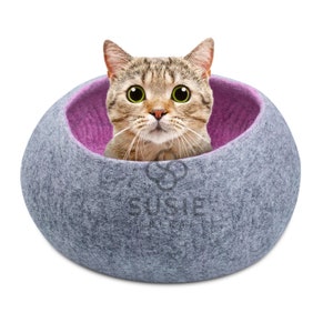 以下が含まれることがあります： グレーとピンクのフェルト製の猫用ベッドで、猫が穴から顔を出しています。猫用ベッドは丸型で、柔らかくふわふわした質感です。