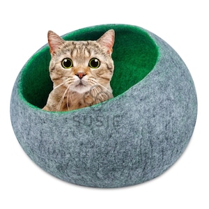 以下が含まれることがあります： 緑色の内装のグレーのフェルト猫用ベッド。大きな緑色の目を 가진トラ猫がベッドの中にいて、視聴者を直接見ています。ベッドは丸みを帯びたポッドのような形をしています。