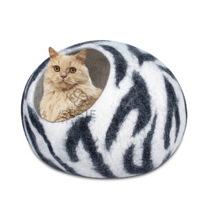 以下が含まれることがあります： 白と黒の模様のフェルト製の猫用ベッド。薄い茶色の猫がベッドの中に座っています。