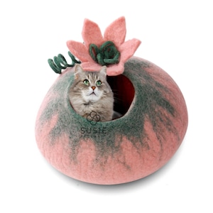 以下が含まれることがあります： 花の形をしたピンクとグリーンのフェルト製の猫用ベッドに猫が入っています。ベッドの上には緑の花が咲き、側面には緑色の渦巻き模様があります。