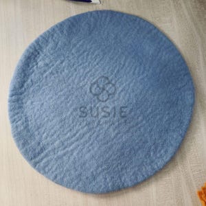 Può includere: Un tappetino rotondo in feltro blu chiaro con una superficie strutturata. Il tappetino presenta il testo "SUSIE EDELCRAFT" in bianco. Il tappetino è di colore pieno e sembra fatto a mano.