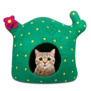 以下が含まれることがあります： 黄色い水玉模様とピンクの花が付いた、サボテンの形をした緑色のフェルト製の猫用ハウス。トラ猫が家の中にいて、正面を見ています。「SUSIE PET」の文字が家の側面に表示されています。