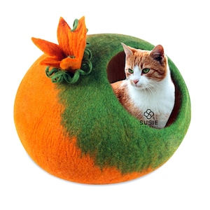 以下が含まれることがあります： 緑とオレンジのフェルト製の猫用ベッドで、上に花が飾られています。猫がベッドの中にいて外を見ています。