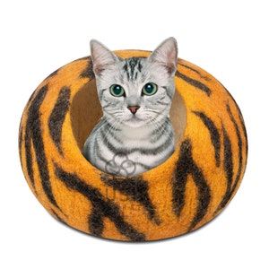以下が含まれることがあります： グレーと白の虎柄の猫が、オレンジと黒の丸い猫用ベッドの中に座っています。ベッドはフェルトでできており、虎の縞模様になっています。