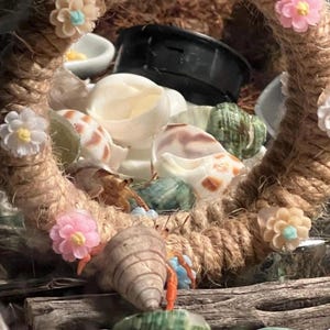 Pu&ograve; includere: Una cornice circolare realizzata in corda spessa e intrecciata color marrone chiaro, decorata con piccoli fiori di plastica colorati nelle tonalit&agrave; del rosa, bianco e pesca. All'interno della cornice ci sono diversi paguri con conchiglie.