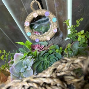 Pu&ograve; includere: Uno specchio circolare incorniciato in corda di juta color cuoio, decorato con piccoli fiori rosa, viola e azzurri. Lo specchio &egrave; appeso sopra una collezione di piante succulente artificiali verdi e viola, con una trama di cesto intrecciato in primo piano.