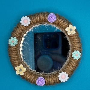 Pu&ograve; includere: Uno specchio circolare con una cornice avvolta in una spessa corda di juta marrone. La cornice &egrave; decorata con otto piccoli fiori di plastica colorati nelle tonalit&agrave; del bianco, viola chiaro e azzurro, disposti uniformemente lungo il bordo.