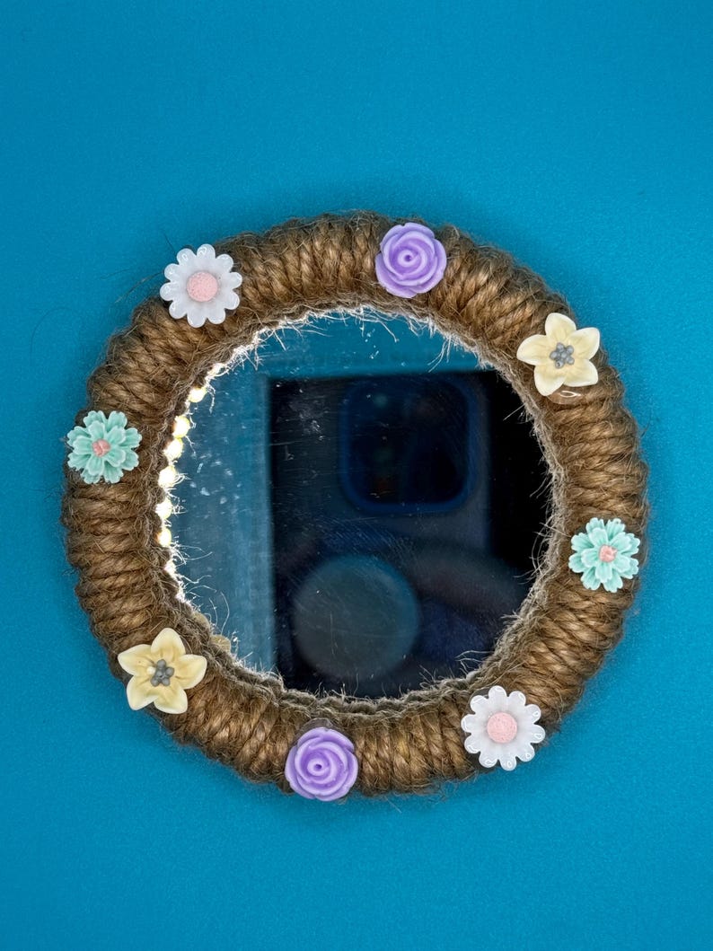 Pu&ograve; includere: Uno specchio circolare con una cornice avvolta in una spessa corda di juta marrone. La cornice &egrave; decorata con otto piccoli fiori di plastica colorati nelle tonalit&agrave; del bianco, viola chiaro e azzurro, disposti uniformemente lungo il bordo.