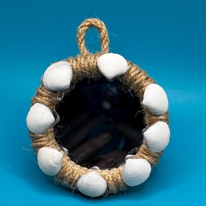 Pu&ograve; includere: Uno specchio circolare incorniciato da una spessa corda di juta color naturale. La cornice &egrave; decorata con otto conchiglie bianche e lisce, distanziate uniformemente lungo il bordo. Un piccolo occhiello di corda &egrave; fissato in alto per appenderlo.