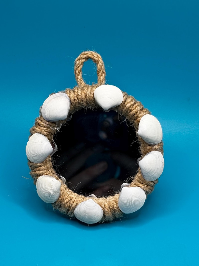 Pu&ograve; includere: Uno specchio circolare incorniciato da una spessa corda di juta color naturale. La cornice &egrave; decorata con otto conchiglie bianche e lisce, distanziate uniformemente lungo il bordo. Un piccolo occhiello di corda &egrave; fissato in alto per appenderlo.