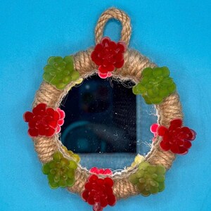 Pu&ograve; includere: Uno specchio circolare con cornice rivestita in corda di juta color marrone chiaro, dotato di un occhiello per appenderlo in alto. La cornice &egrave; decorata con ornamenti a forma di piante succulente rosse e verdi alternate lungo il bordo.