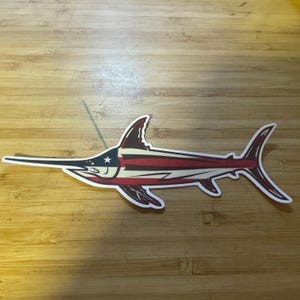 May include: A sticker of a swordfish with a patriotic design. The body of the fish features red and white stripes, with a blue section containing a white star. The sticker has a white border and is on a wooden surface.