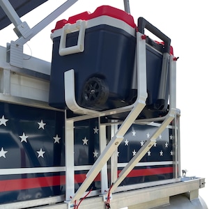 May include: A white metal mounting rack attached to the side of a boat, holding a large navy blue, white, and red cooler with wheels. The boat exterior features a navy blue panel with white stars and a horizontal red stripe along the bottom.