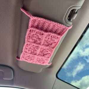 Könnte beinhalten: Ein hellrosa gehäkelter Auto-Sonnenblenden-Organizer mit einem strukturierten Sternmuster. Das rechteckige Zubehör ist aus Garn gefertigt und verfügt über eine obere Öffnung, die so konzipiert ist, dass sie sicher über eine Sonnenblende passt.