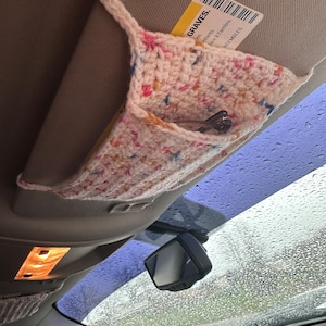 Puede incluir: Un organizador de visera de coche de ganchillo blanco roto hecho a mano con motas de colores. El organizador tiene un bolsillo que contiene un ticket de aparcamiento y un juego de llaves. La visera está sujeta al interior del coche.