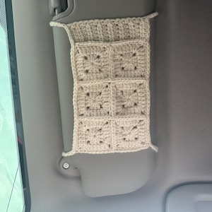 May include: A cream-colored, crocheted car visor organizer. The organizer is attached to a gray car visor and features a series of square pockets. The crochet work has a textured, open design, and the overall aesthetic is handmade and rustic.