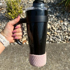 Puede incluir: Una taza de viaje de acero inoxidable negro con un asa negra y un acogedor de crochet rosa. La taza tiene una tapa transparente con una pajita negra.