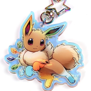 Puede incluir: Un charm holográfico con un dibujo animado de Eevee sobre un fondo azul, verde y naranja. El charm está unido a un llavero en forma de estrella plateada.
