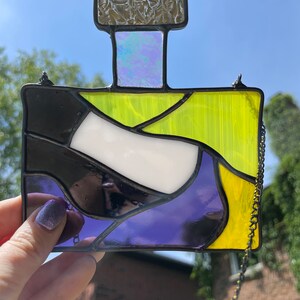 Könnte beinhalten: Ein Buntglasfenster, das an einer Kette hängt. Das Fenster ist in Form einer Parfümflasche gestaltet und zeigt ein Design aus schwarzem, weißem, gelbem und violettem Glas.