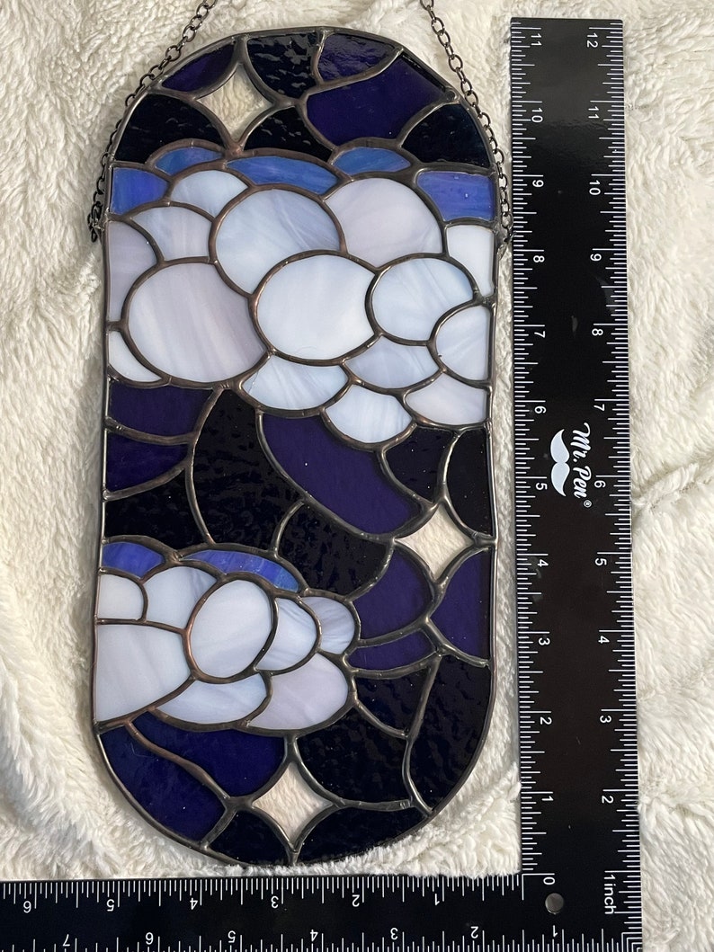 May include: A stained glass window hanging with a chain. The window is oval shaped and features a design of white and blue flowers on a dark blue background.