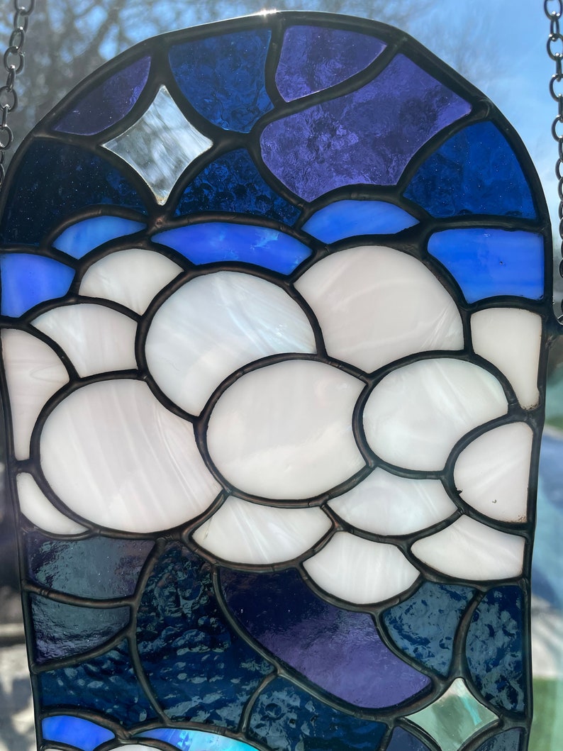 May include: A stained glass window hanging with a blue, purple, and white design. The design features a cloud-like shape with a star in the upper left corner.