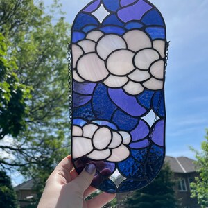 May include: A stained glass window hanging with a chain. The window is oval shaped and features a blue and purple background with white flowers.