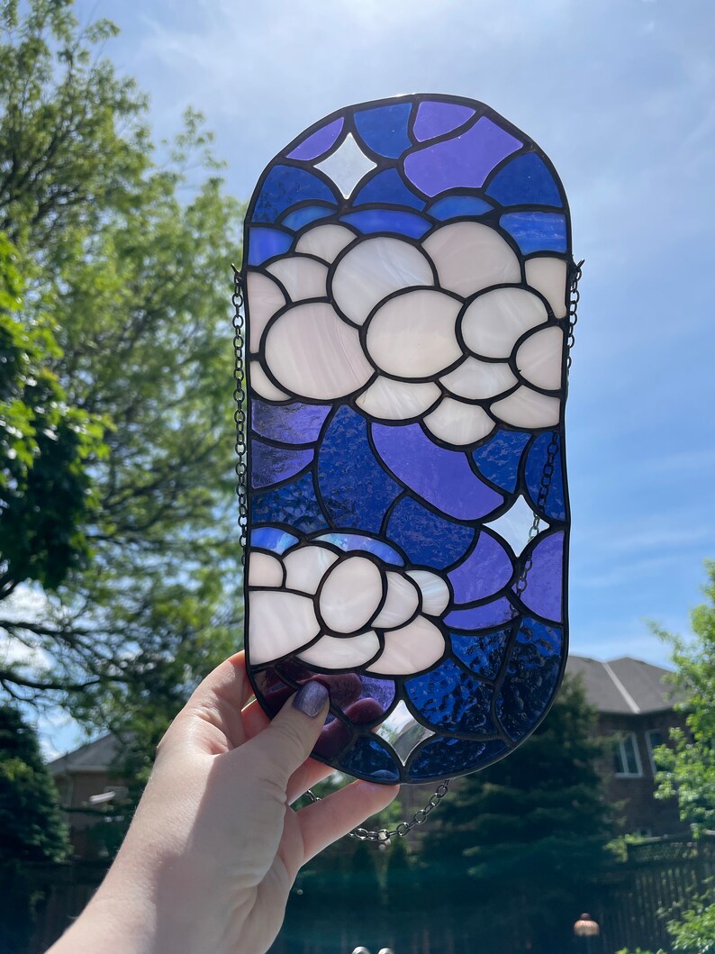 May include: A stained glass window hanging with a chain. The window is oval shaped and features a blue and purple background with white flowers.