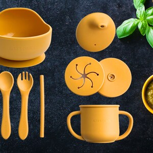 May include: A set of yellow silicone baby feeding products, including a bowl with a suction base, a cup with two handles, a spoon, a fork, a straw, and two lids. The lids have a star-shaped design and a plus sign.