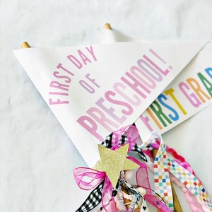 Op de afbeelding: Een witte vlag met de tekst "First Day of Preschool!" in roze en paarse letters. De vlag is versierd met roze, zwart-wit geblokt lint, roze lint en een gouden ster.