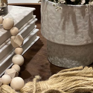 Puede incluir: Una guirnalda decorativa de cuentas de madera con cuentas geométricas y redondas, apoyada sobre una pila de libros blancos. Una maceta de cerámica gris claro texturizada con flores blancas se encuentra cerca en una bandeja de madera. También hay un manojo de hilo natural.
