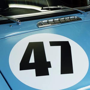 Peut inclure: Un capot de voiture bleu clair arborant un grand autocollant circulaire blanc avec le numéro 47 imprimé en noir au centre. Le capot comprend une grille d'aération en métal argenté et deux essuie-glaces argentés près de la base du pare-brise.