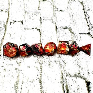 May include: A set of seven polyhedral dice, each with a unique shape, arranged in a row. The dice are transparent with a red and gold color scheme, and black numbering. The dice are designed for tabletop role-playing games.
