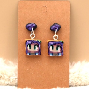 May include: A pair of dangle earrings featuring a pixelated character with purple hair and a blue shirt. The character is surrounded by a brown frame, resembling a slice of toast.