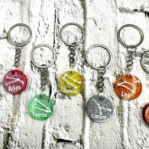 May include: Keychains featuring zodiac signs: Aries, Gemini, Taurus, Leo, and Cancer. Each keychain has a round, glittery charm with a constellation design and sign name in white. The charms are attached to a silver chain and keyring.