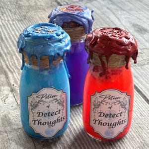 May include: Three glass bottles filled with blue, purple, and red liquid. Each bottle has a label reading Potion of Detect Thoughts, a twine-wrapped neck, and a wax seal on the cork featuring star and moon designs with dripping wax edges.