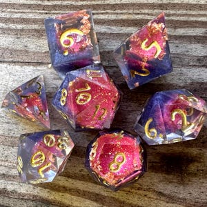 May include: A set of seven polyhedral dice made of translucent resin with a swirling purple and pink interior, embedded with gold leaf flakes. Each die features engraved numbers filled with gold paint, including a d20, d12, d10, d8, d6, and d4.
