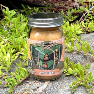 May include: A clear glass jar with a silver metal lid containing a light orange candle. The label features a green, square-shaped loaf tied with string and the text ELVEN BREAD printed in white on a light brown rectangular background.