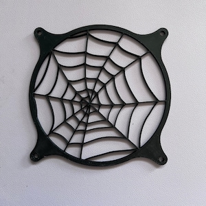 May include: A black, circular fan grill featuring a spiderweb design. The grill has four mounting tabs and a detailed web pattern, perfect for Halloween or gothic decor. The item is made of a solid, dark material.
