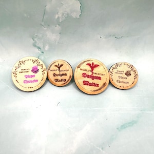 May include: Four circular wooden tokens with engraved text and designs. Two tokens read World's Greatest Player Character with a small icon, and two tokens read World's Greatest Dungeon Master with a dragon icon and pink or brown lettering.