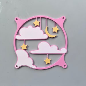 May include: A pink, circular decorative piece featuring white clouds, yellow stars, and a crescent moon. The design is suspended within a pink frame with four small holes, creating a whimsical, celestial theme. Ideal for nursery decor.