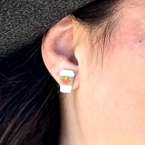 May include: A white and green coffee cup stud earring with a pumpkin design. The earring is worn on the ear of a person. The earring is a miniature version of a coffee cup, with a white base and a green and orange design.