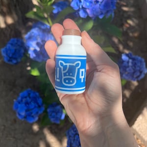 May include: A small, white bottle with a brown cap, featuring a blue label with a cow illustration. The bottle is held in a hand, with blue flowers in the background. The label has the word "milk" in Japanese.