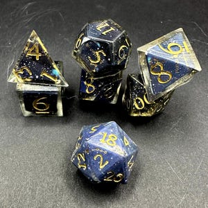 May include: A set of polyhedral dice for tabletop games. The dice are a deep blue with gold numbering and a starry night effect. The set includes a D4, D6, D8, D10, D12, and D20.