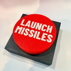 May include: A circular red button with the white text LAUNCH MISSILES printed on the top surface. The button is mounted on a square black base with a textured finish. The design features a bold, high-contrast aesthetic with clear lettering.
