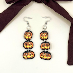 Puede incluir: Un par de pendientes colgantes con tres abalorios de calabaza tallada en forma de jack-o'-lantern apilados. Cada abalorio es una calabaza negra y naranja con una cara sonriente tallada.