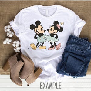 K&ouml;nnte beinhalten: Wei&szlig;es T-Shirt mit einer Cartoon-Illustration von Mickey und Minnie Mouse. Mickey tr&auml;gt hellblaue Shorts und gelbe Schuhe, Minnie ein gepunktetes Kleid und rosa Schuhe. Das Wort "EXAMPLE" ist unten aufgedruckt.