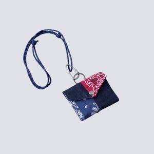 Puede incluir: Una pequeña bolsa rectangular hecha de mezclilla azul oscuro y tela estampada con diseños de cachemira en rojo y azul. La bolsa tiene un cierre de broche plateado, un anillo de metal, un mosquetón plateado y un cordón azul con puntos blancos.