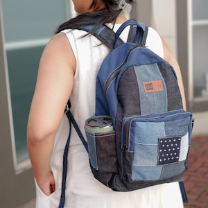 May include: A patchwork backpack constructed from various shades of blue denim fabric. The bag features a front zippered pocket with a small square patch displaying a white cross pattern, a side pocket holding a tumbler, and a rectangular brand label.