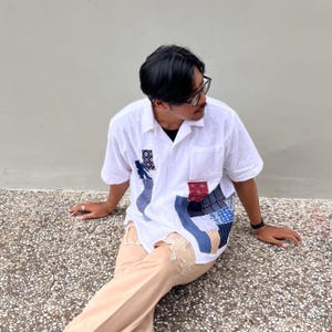 Pode incluir: Uma camisa branca de manga curta com botões e um design de patchwork assimétrico na frente. Os remendos consistem em vários quadrados de tecido azul e vermelho com padrões geométricos, combinados com calças de cor castanho-claro.